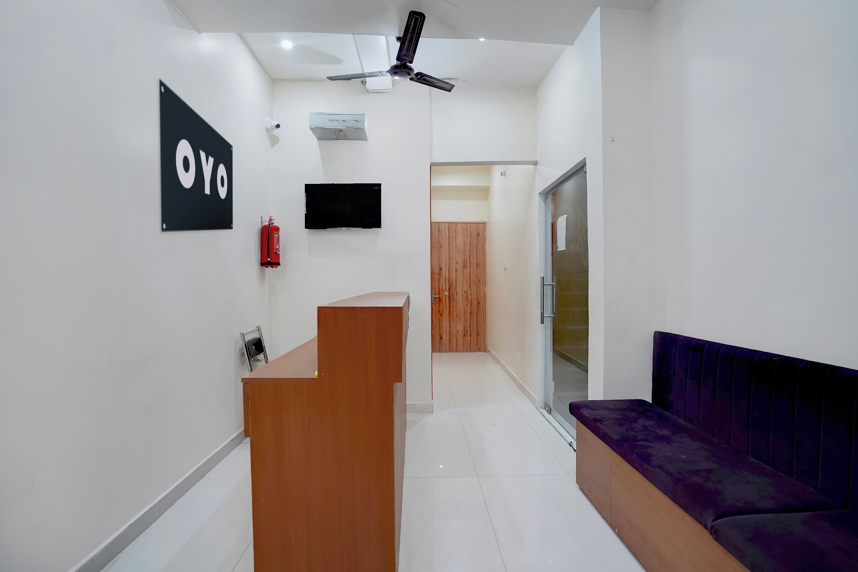 Una zona de recepción con un escritorio de madera, un letrero negro, un extintor, un ventilador de techo y un sofá morado.