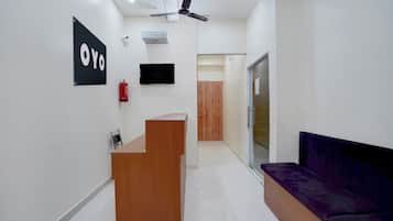 Una zona de recepción con un escritorio de madera, un letrero negro, un extintor, un ventilador de techo y un sofá morado.