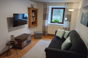 Ein Wohnzimmer mit Flachbildfernseher, Holzschrank, schwarzem Sofa mit Kissen und kleinem Tisch mit Fernbedienung. Ein Fenster mit Vorhängen ist ebenfalls zu sehen.