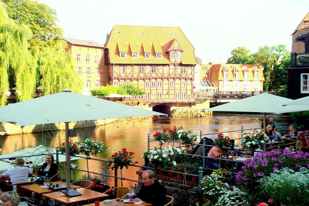 Un espace de restauration au bord de la rivière, avec des tables et des parasols, un pont et des bâtiments traditionnels en arrière-plan.