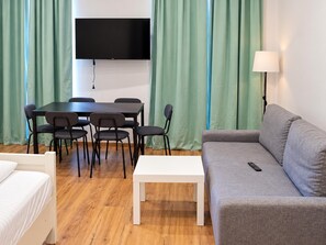 Een kamer met een wandgemonteerde flatscreen-tv, een eettafel met stoelen, een bank, een witte bijzettafel en een lamp.