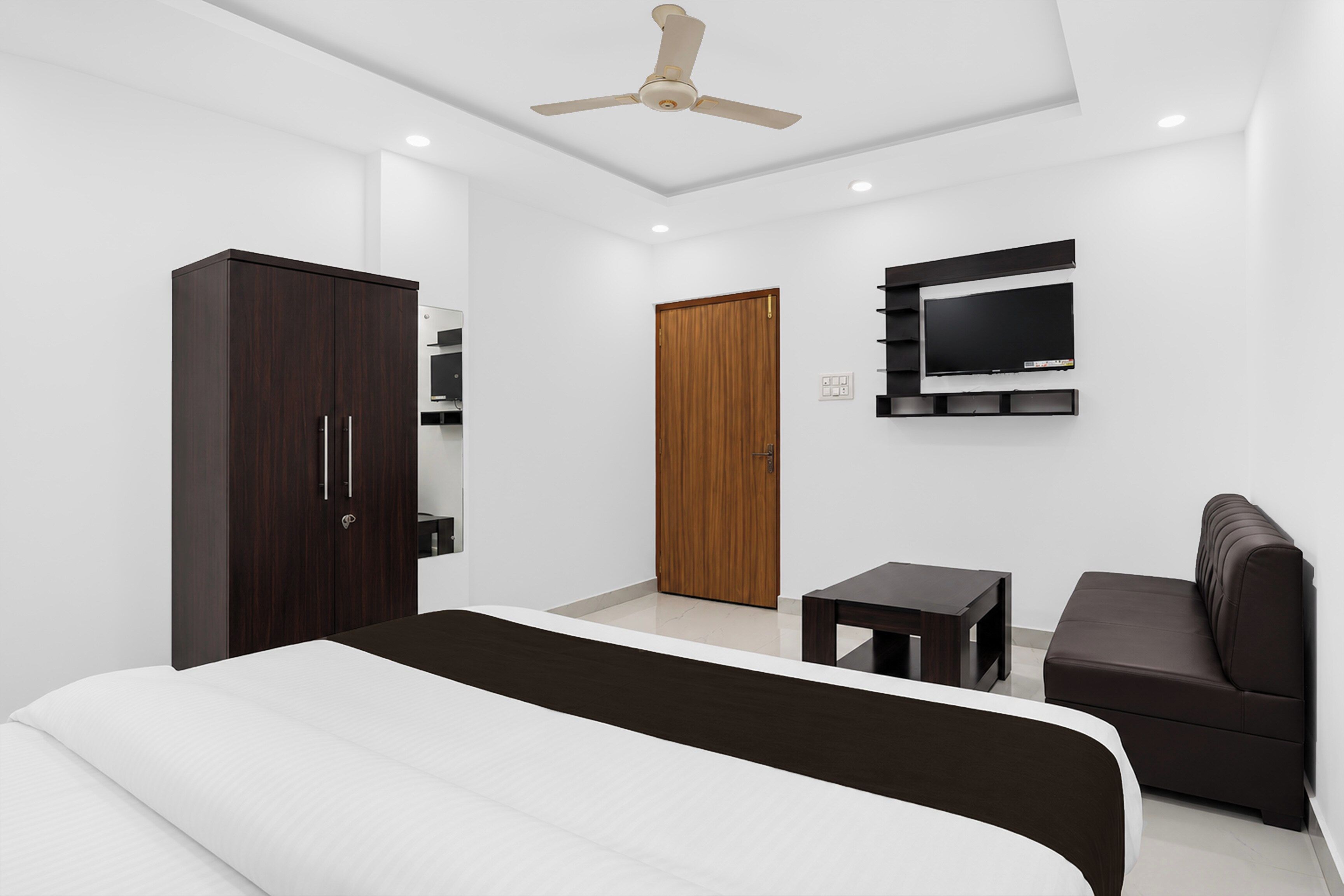 A modern bedroom with a bed, a wardrobe, a television mounted on the wall, a ceiling fan, and a small table.