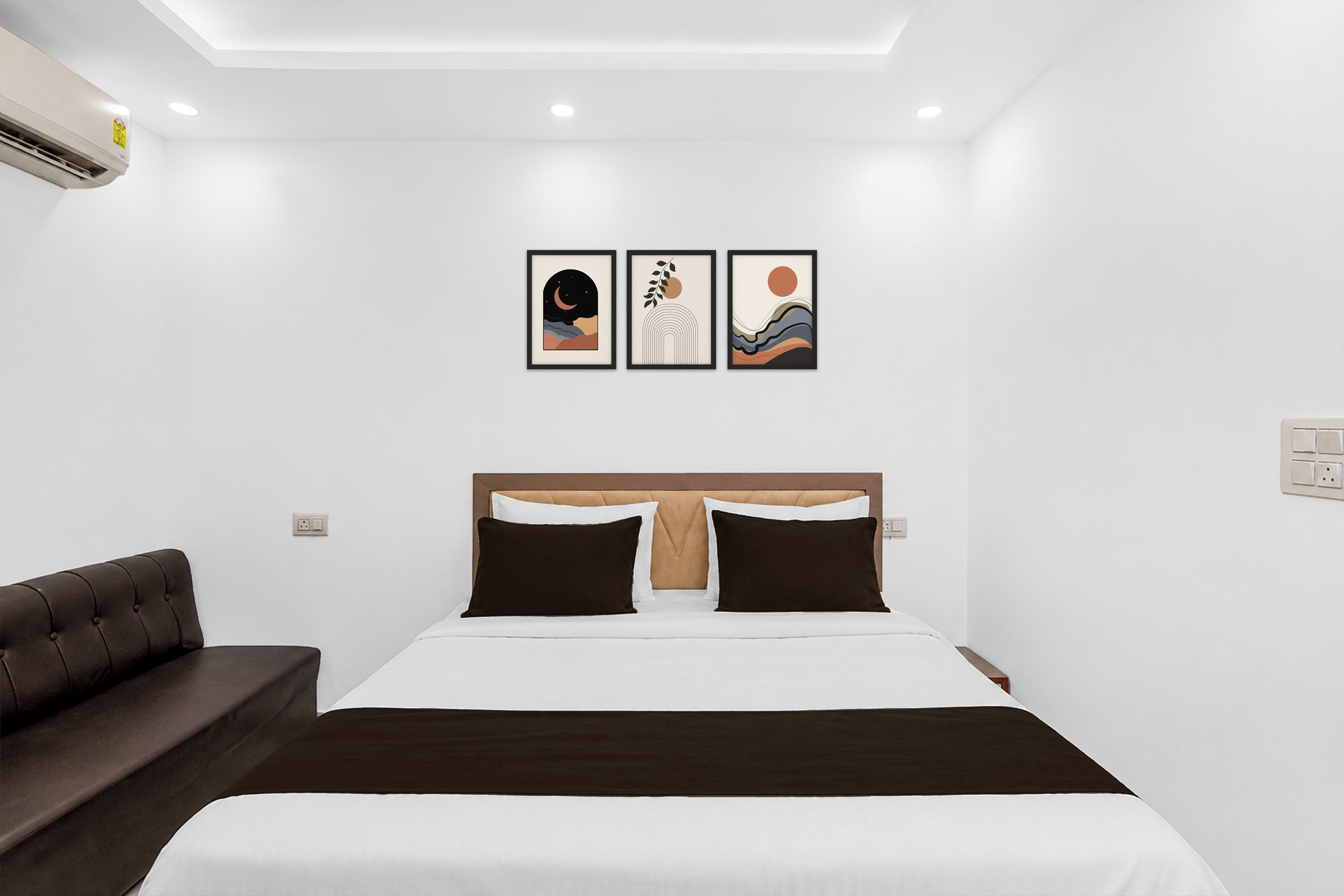 A hotel room with a bed, a brown sofa, and three framed artworks on the wall.