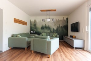 A modern living room with a green sofa set, a flat-screen TV, and a forest mural on the wall. - Holiday home South Harz (Neustadt\/Harz)