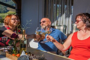 Eine Gruppe von Menschen, die mit Weingläsern anlächeln, ein Tisch im Freien mit einer Weinflasche und einer Glasvase mit einer Blume.