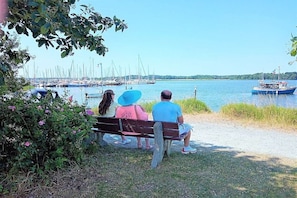 A bench with three people sitting, overlooking a body of water with boats and a dock in the background. - Holiday apartment with 1 bedroom (Maasholm)