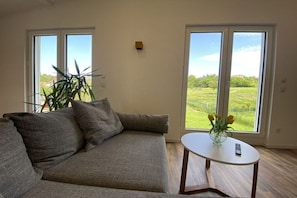 A living room with a grey sofa, a small white table, and a view of a green landscape through large windows. - Holiday home with 2 bedrooms (Nohfelden OT Eckelhausen)