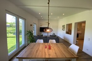 A modern interior with a wooden dining table, white chairs, a hanging light fixture, and a view of the outdoors through large windows. - Holiday home with 2 bedrooms (Nohfelden OT Eckelhausen)