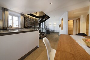 A modern kitchen with a central island, a staircase with a stone accent wall, and a dining area with a wooden table. - Heimat1883 - Coachman (Garmisch-Partenkirchen)