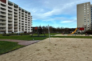 Un terrain de jeux avec des balançoires et un toboggan, entouré d’une pelouse et bordé d’un bâtiment à plusieurs étages.