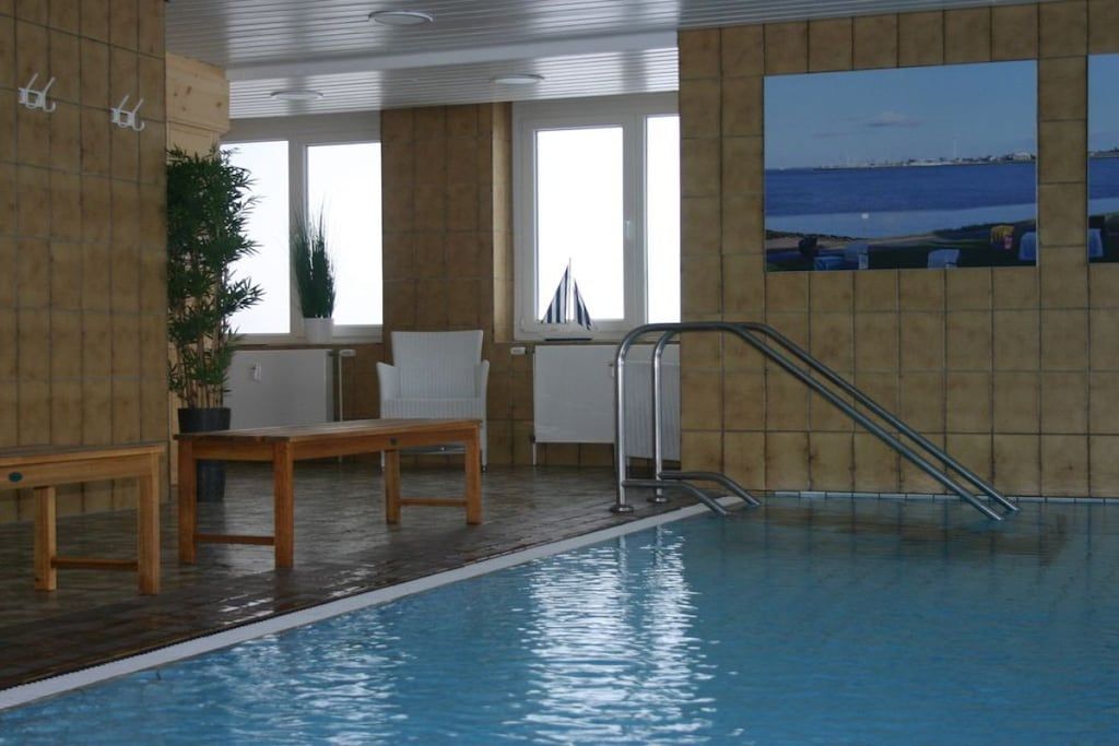 An indoor swimming pool with a wooden bench, a chair, and a view of the sea through large windows.