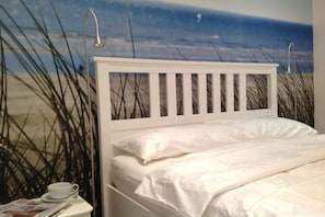 Un lit avec une tête de lit blanche, une tasse et une soucoupe sur une table de chevet, et un mur décoré d’une scène de plage.