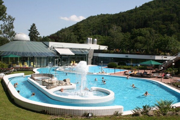 A swimming pool with a fountain, surrounded by lounge chairs and a building with a green roof. - Goldberg Forest Lodge (Bad Harzburg)