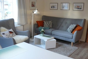Un salon avec un canapé gris, une table basse blanche et un fauteuil bleu. Deux tableaux encadrés sont accrochés au mur, et une lampe sur pied est posée au sol.