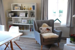 A living room with a grey armchair, a white bookshelf, and a window with curtains. - Holiday apartment with 1 bedroom (Rendsburg)