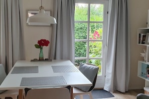 A dining area with a white table, grey cushions, a vase with a red flower, and a window with curtains. - Holiday apartment with 1 bedroom (Rendsburg)
