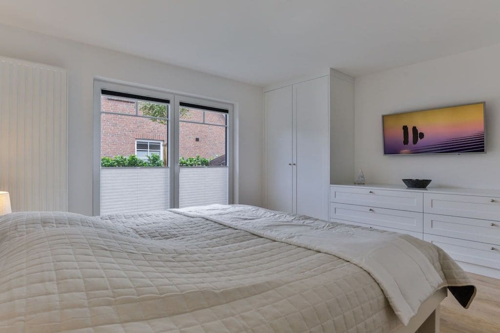 Une chambre à coucher avec un lit, une commode, un téléviseur et une fenêtre donnant sur un immeuble en briques.