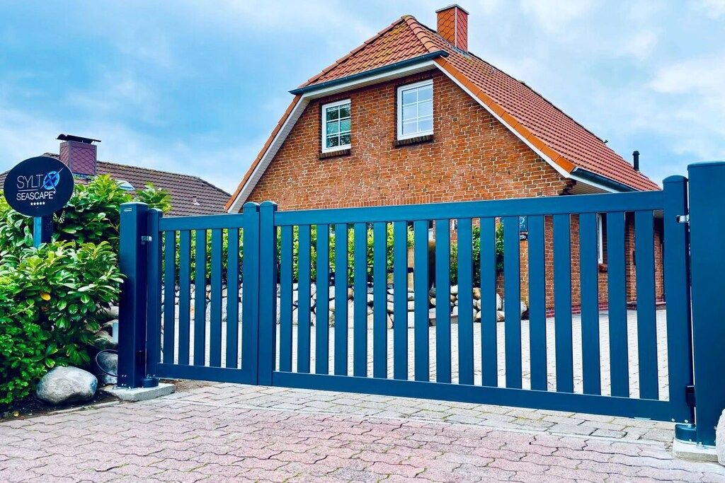 A brick house with a blue gate, a sign with 'SYLT' and 'SEA SCAPE' visible, and a paved driveway.