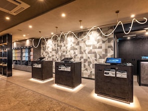 A modern reception area with a patterned wall, three dark reception desks with printers, and a digital display. - Super Hotel Shiga Nagahama Tennen Onsen (Nagahama)