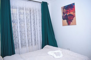 Une chambre à coucher avec un lit, une fenêtre ornée de rideaux légers, un tableau accroché au mur et un oreiller blanc.