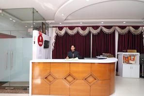 A reception desk with a person seated behind it, a reception area with a red curtain backdrop, and a sign indicating the reception location. - Hotel O Misti Inn (Jamshedpur)