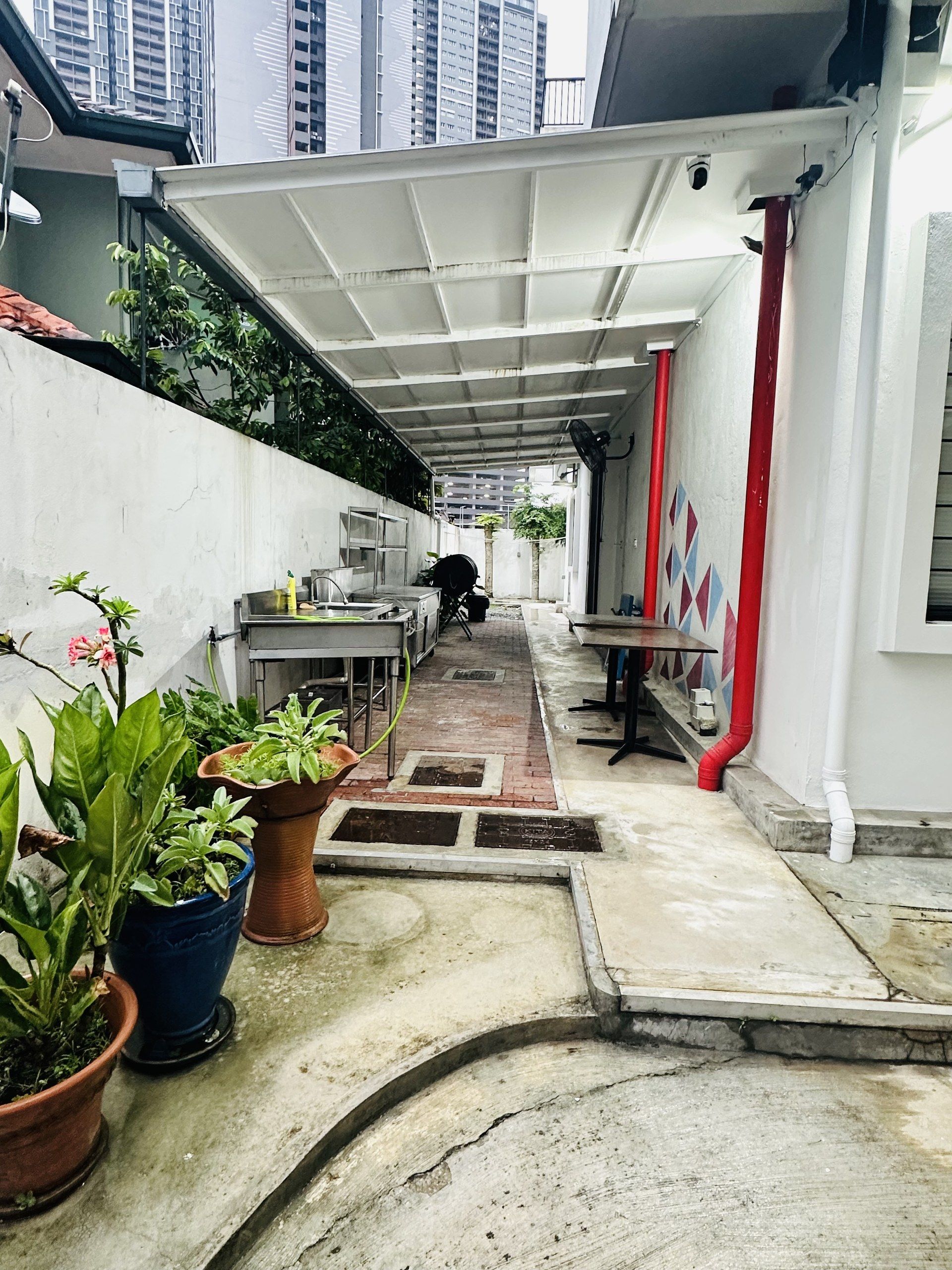 A narrow corridor with a tiled floor, potted plants, a metal table, and a red pipe.