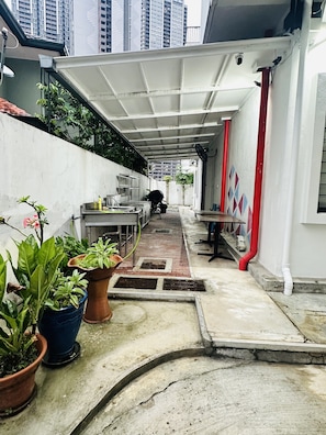 Um corredor estreito com chão de azulejos, plantas em vasos, uma mesa de metal e um cano vermelho.