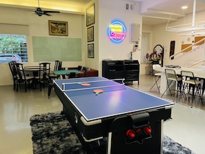 A room with a ping pong table, chairs, a desk, a bookshelf, and a neon sign.