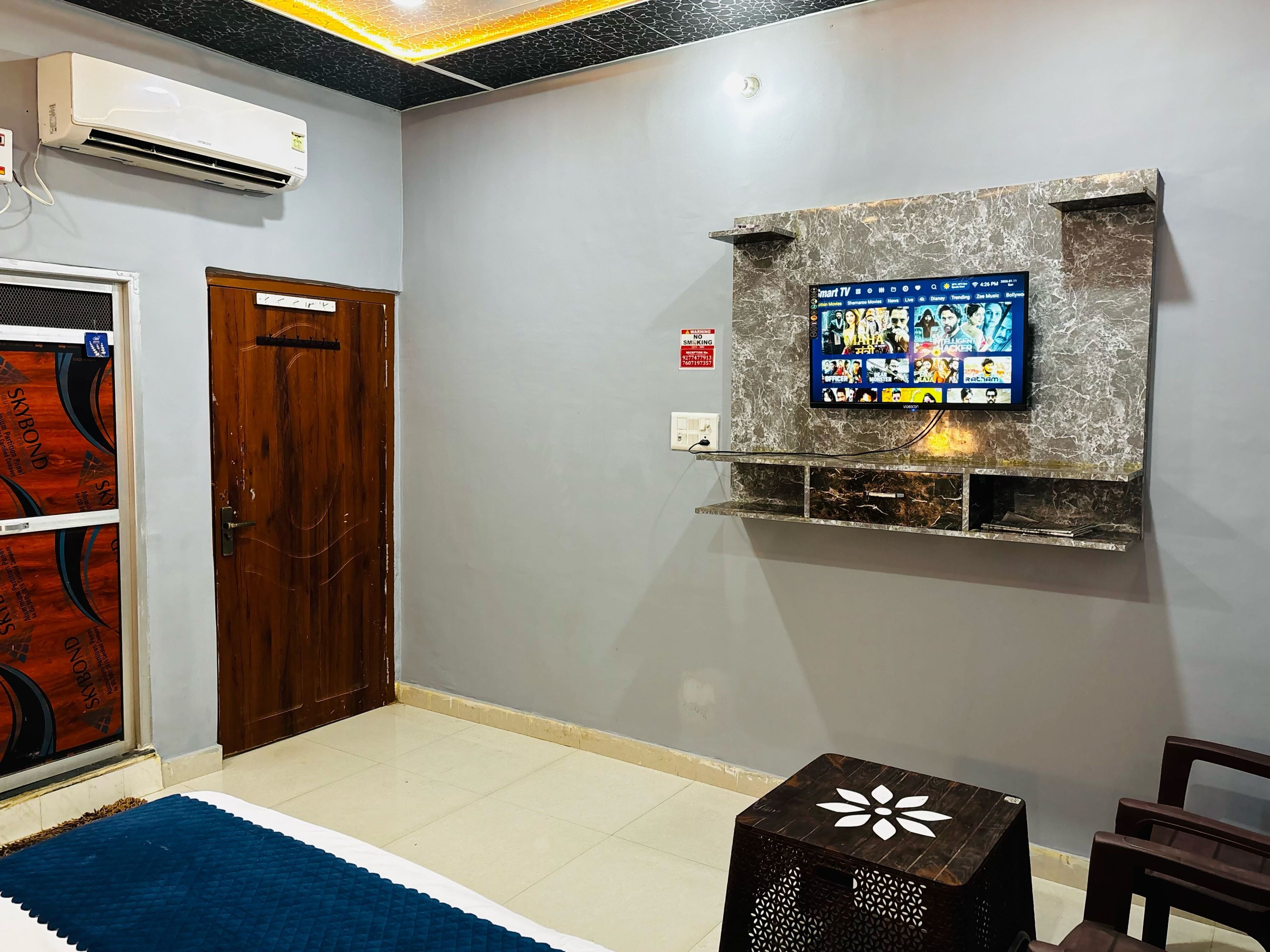A room with a mounted TV on a marble wall, a wooden door, and a small table with a decorative top.
