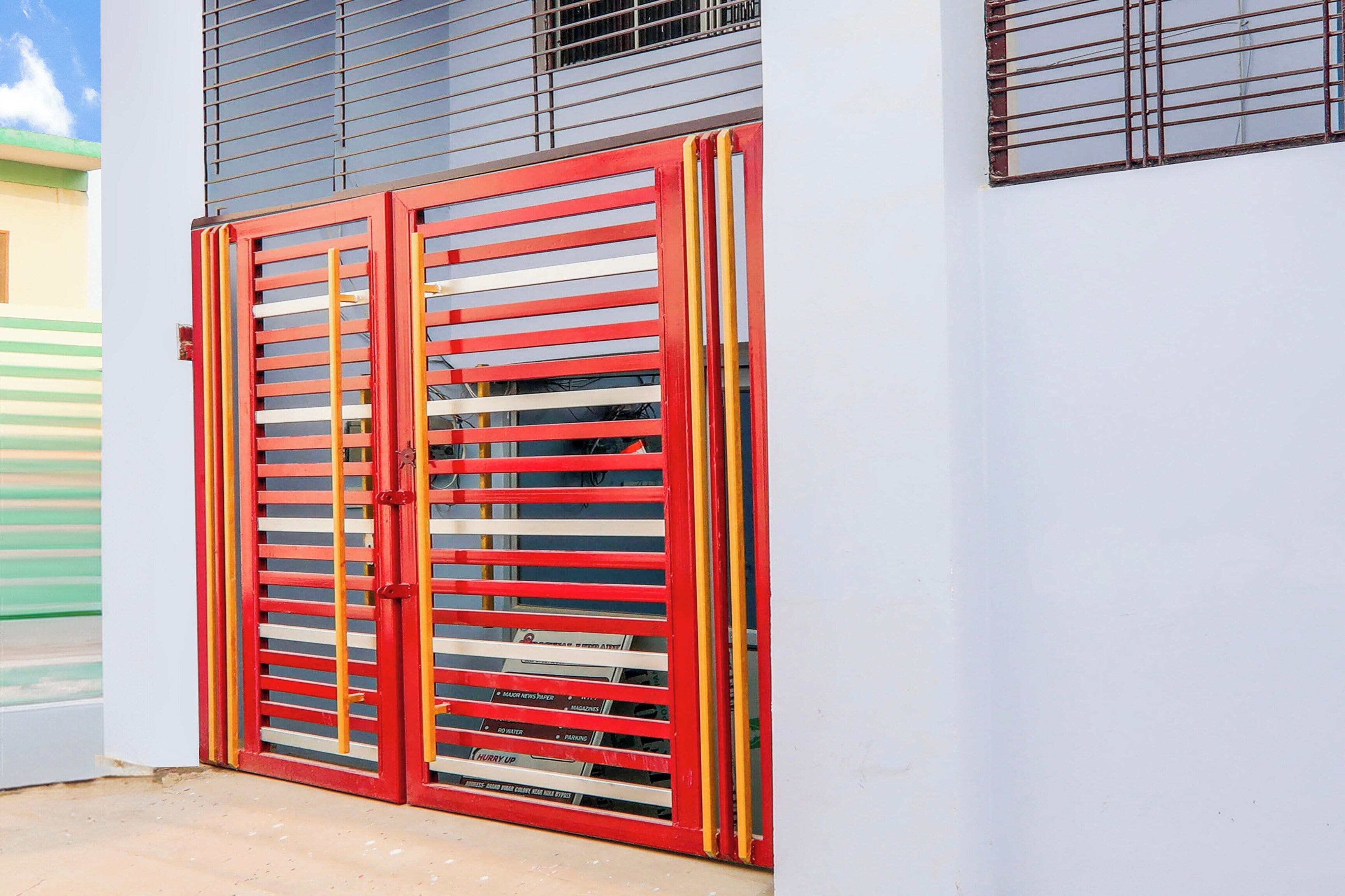 Une porte métallique rouge, avec des barreaux horizontaux, fixée à un bâtiment dont le mur est blanc et le toit bleu.