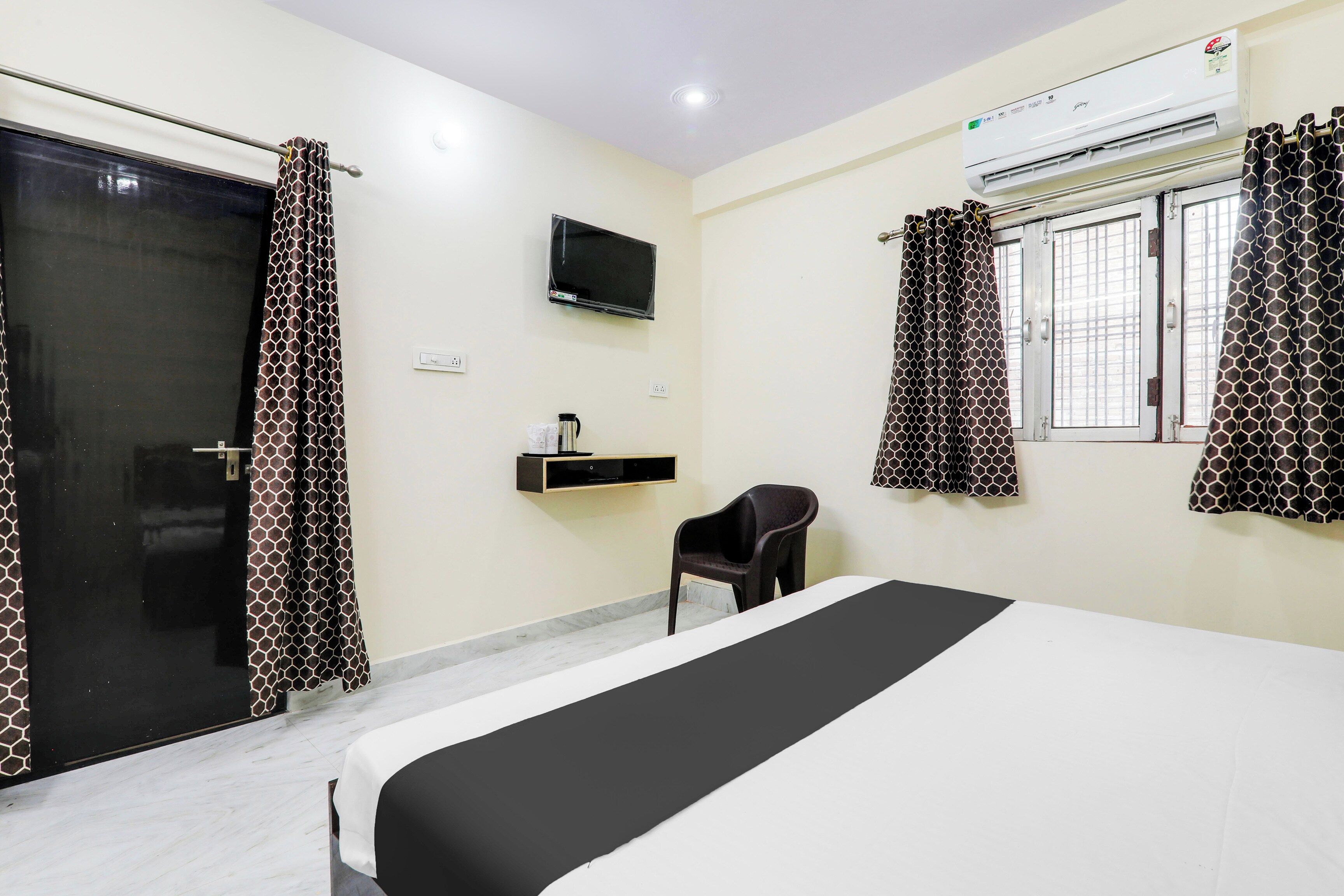 Une chambre d’hôtel avec un lit, un téléviseur fixé au mur, une fenêtre avec des rideaux, une petite table avec une bouilloire et une chaise.