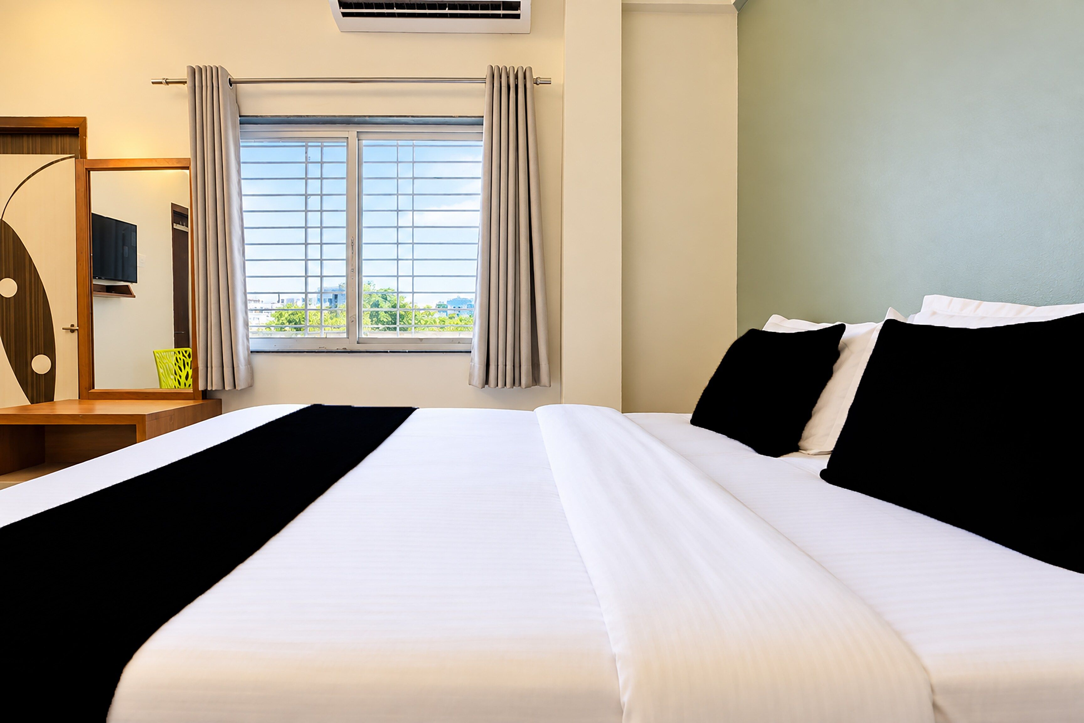Une chambre d’hôtel avec un grand lit, un téléviseur posé sur un meuble en bois, une fenêtre avec des rideaux et une vue sur la verdure à l’extérieur.