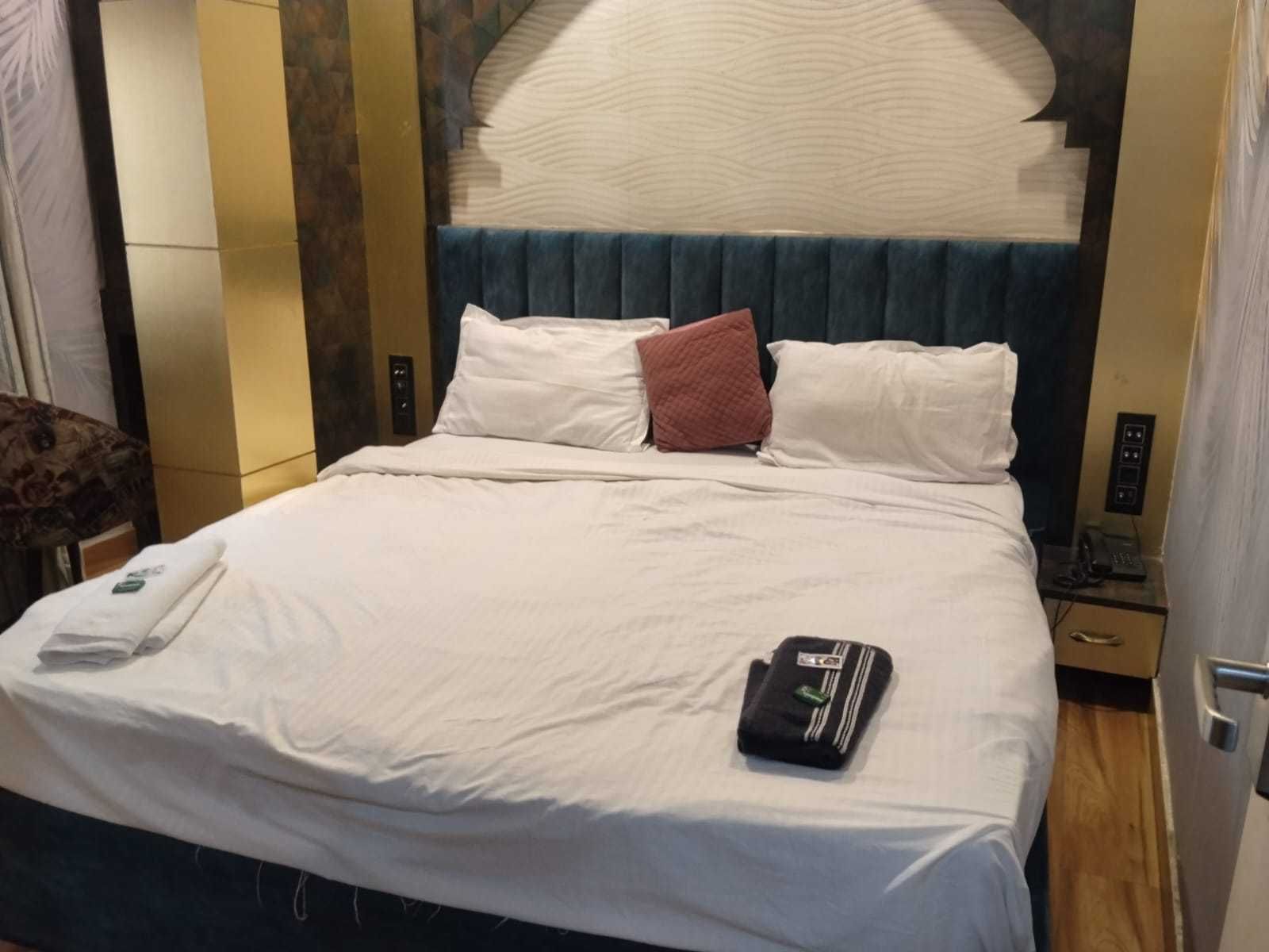 A hotel room with a bed, a bedside table with a phone, and a wall-mounted control panel.