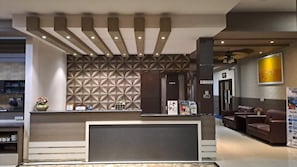 Zona de recepción de hotel con diseño moderno, que incluye una pared con patrón geométrico, mostrador de recepción, área de descanso y un cartel de prohibido fumar visible.