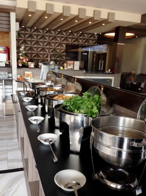 Montaje de bufé con múltiples estaciones de comida, que incluye barra de ensaladas y diversos platos calientes, en un moderno entorno hotelero.