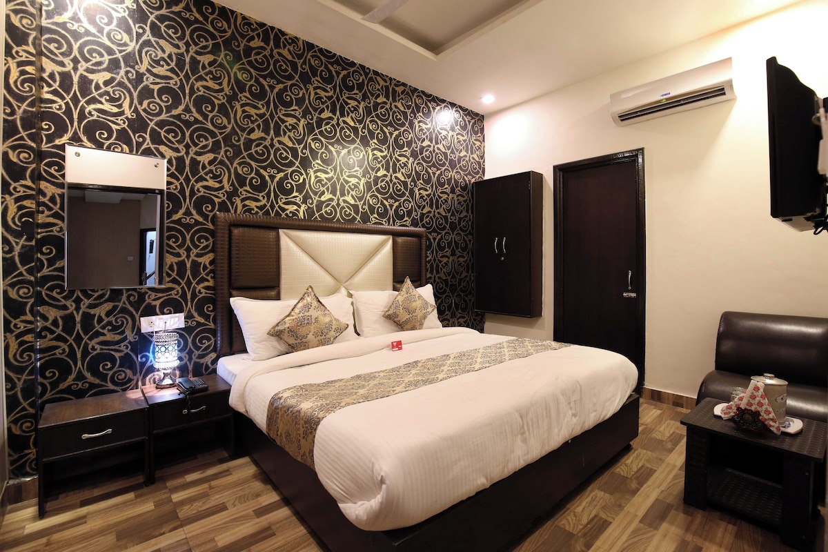 Habitación de hotel con una cama grande, una mesita de noche con lámpara, un armario, un televisor y una zona de estar con mesa y sillón.