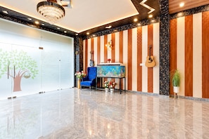 A lobby with a marble floor, a blue chair, a small table with flowers, and a wall with vertical wooden panels and a guitar. - Hotel O Aurivique hotels and Banquets (Lucknow)