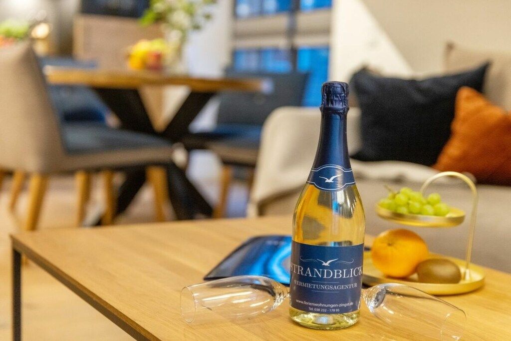 Une bouteille de vin et un verre sur une table en bois dans un salon, avec un canapé et un coin repas en arrière-plan.