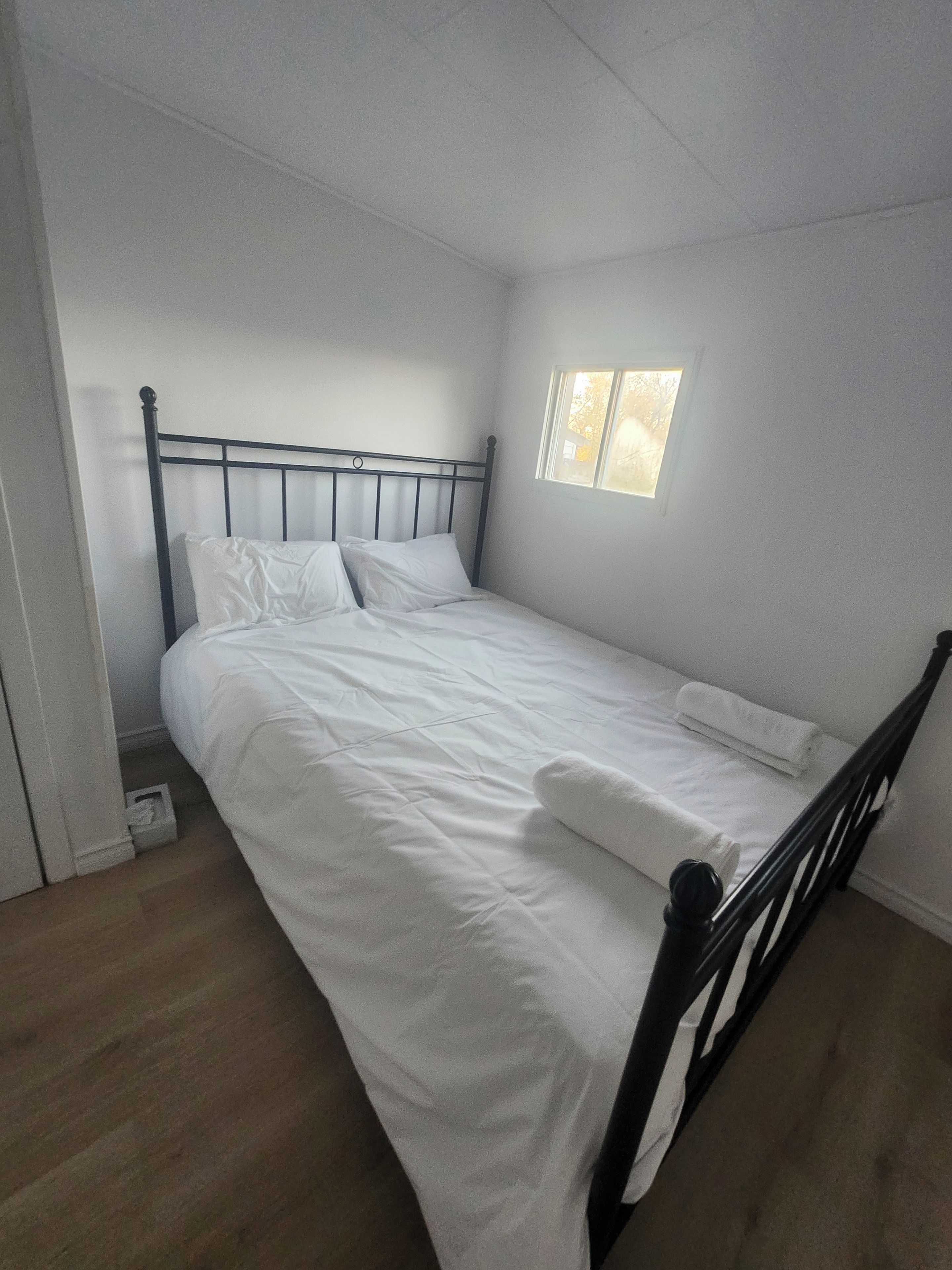 Un lit simple avec un cadre en métal et une literie blanche, dans une pièce dotée d’une petite fenêtre.