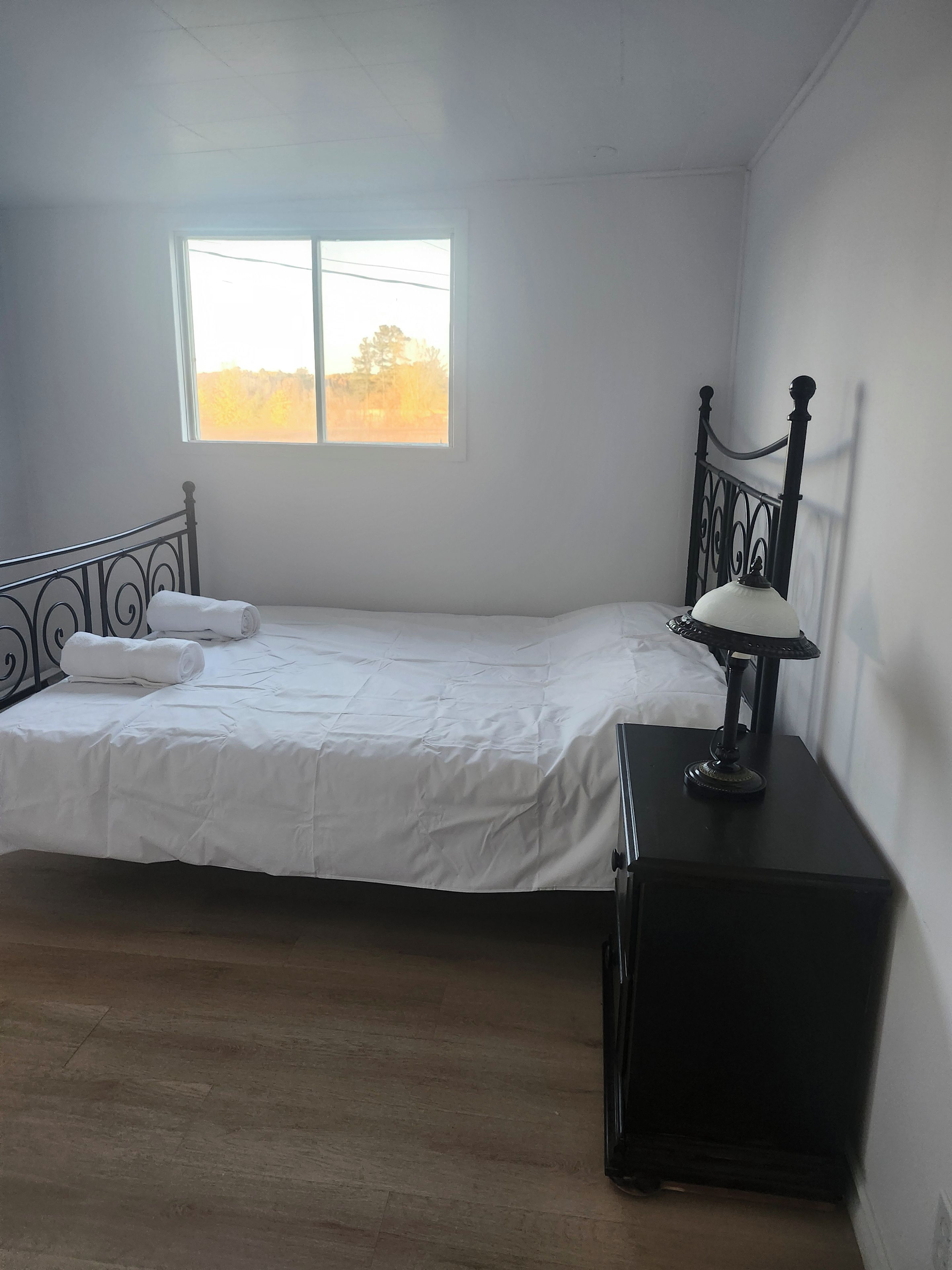Un lit simple avec un matelas blanc et une tête de lit en métal noir, dans une pièce dotée d’une fenêtre.