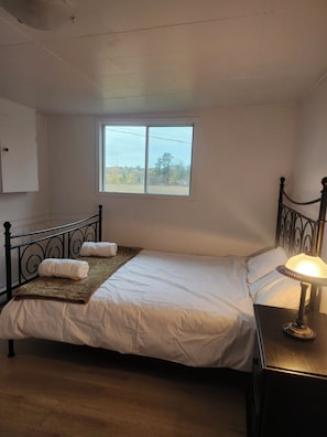 En enkel seng med gavl, et sengebord med lampe, et vindue med udsigt til træer og et vægmonteret skab.
