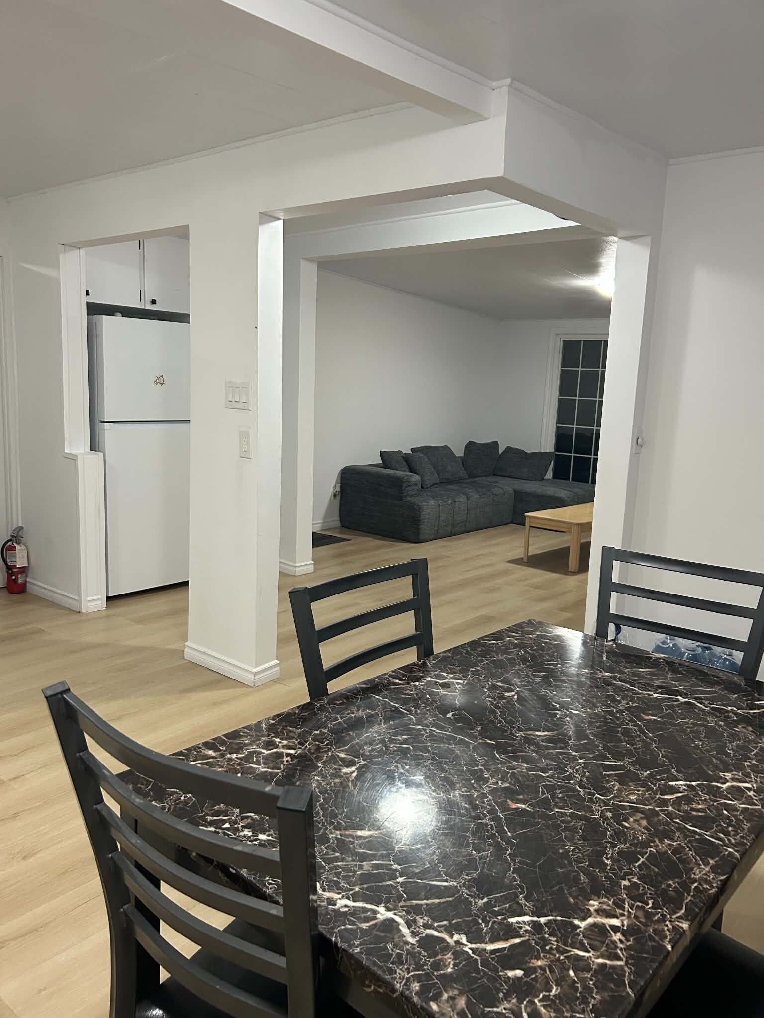 A dining area with a marble table and chairs, a kitchen with white cabinets, and a living room with a grey sofa.