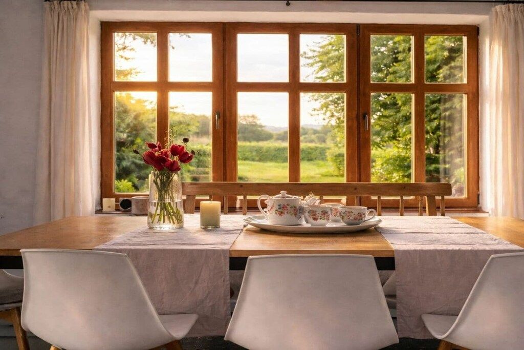 Un comedor con una mesa dispuesta para dos, un florero con flores y vistas a la vegetación a través de amplias ventanas.