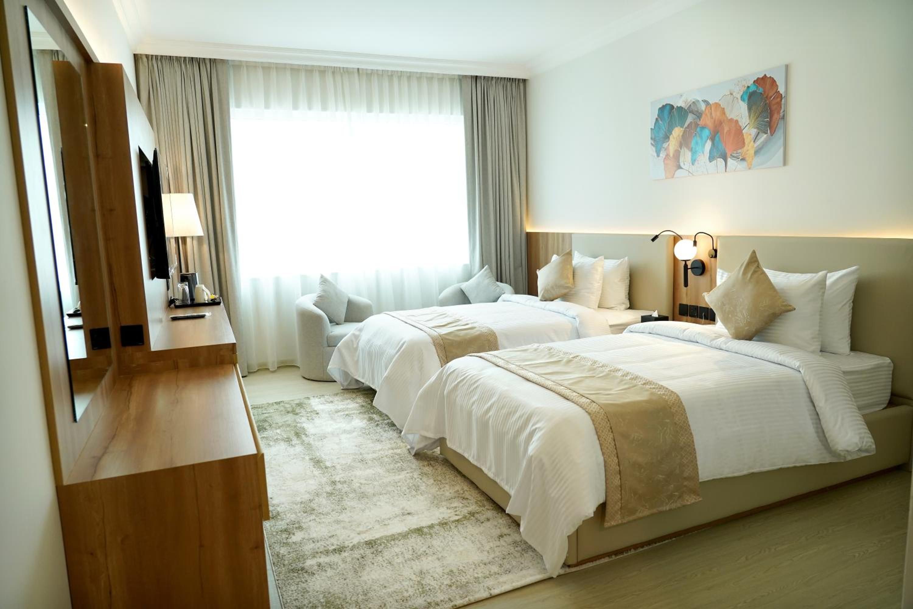 A hotel room with two beds, a desk, a chair, a large window with curtains, and a painting on the wall.
