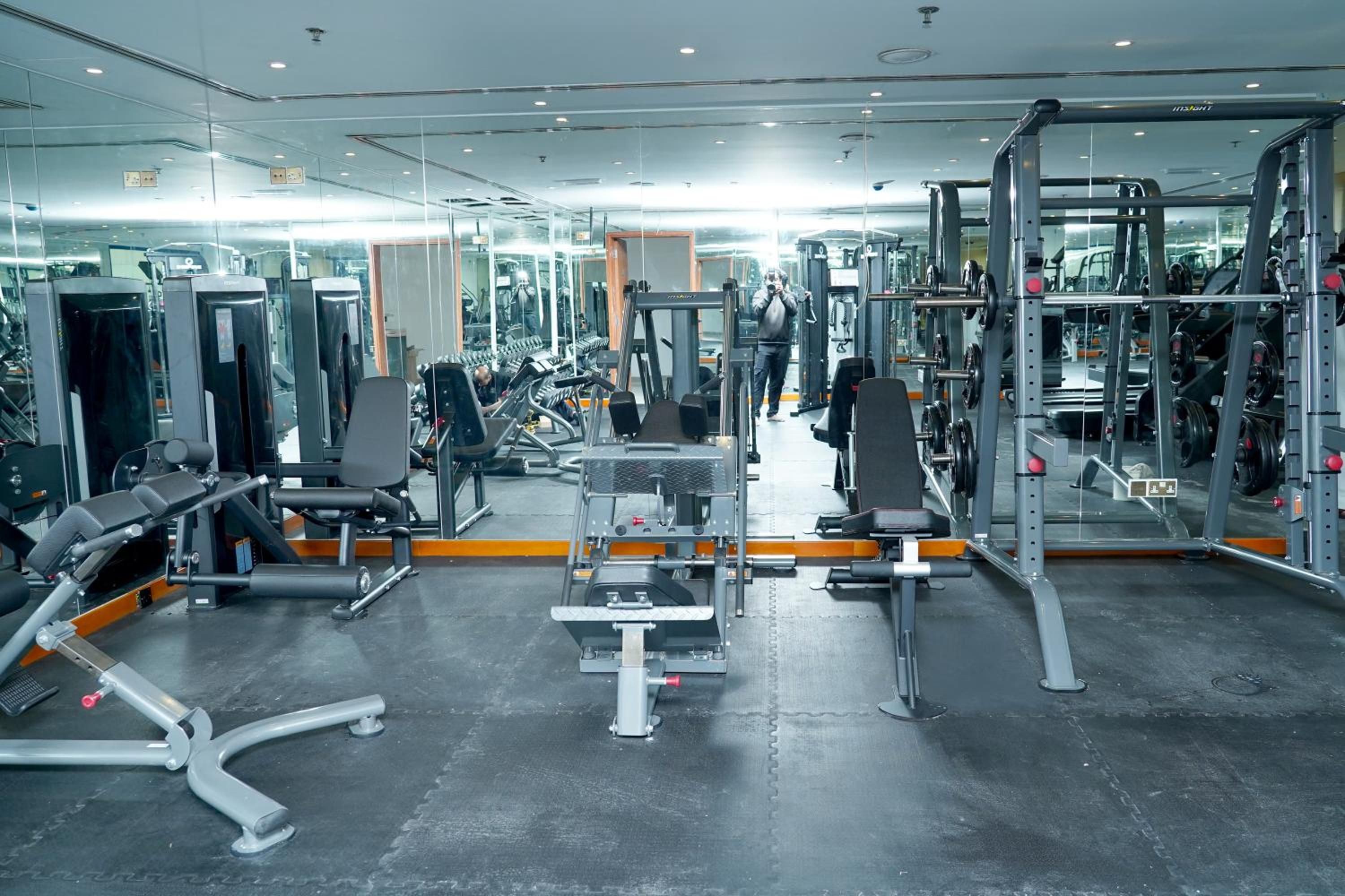 Une salle de sport équipée de divers appareils d’exercice, de miroirs, et où une personne utilise une machine.