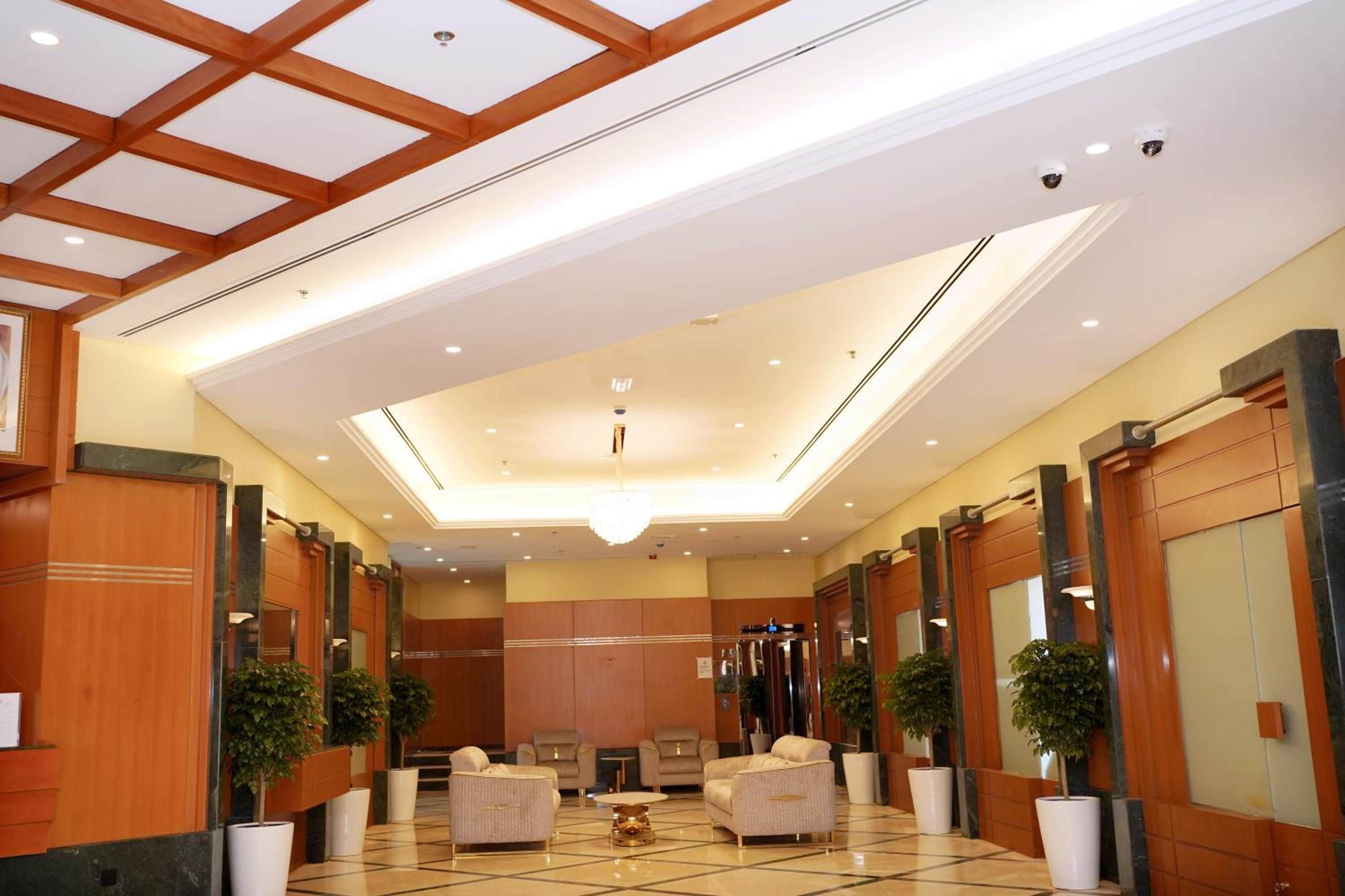 A hotel lobby with a high ceiling, recessed lighting, and a central chandelier. There are seating areas with sofas and armchairs, and potted plants are placed along the walls.