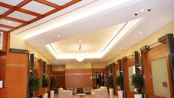 A hotel lobby with a high ceiling, recessed lighting, and a central chandelier. There are seating areas with sofas and armchairs, and potted plants are placed along the walls.