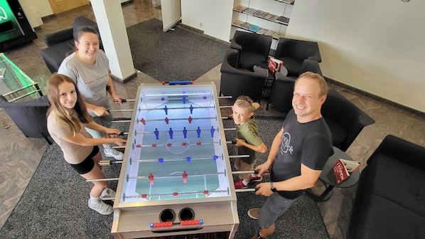 Un grup de persones juga al futbolí en una habitació amb terra de moqueta i cadires negres.