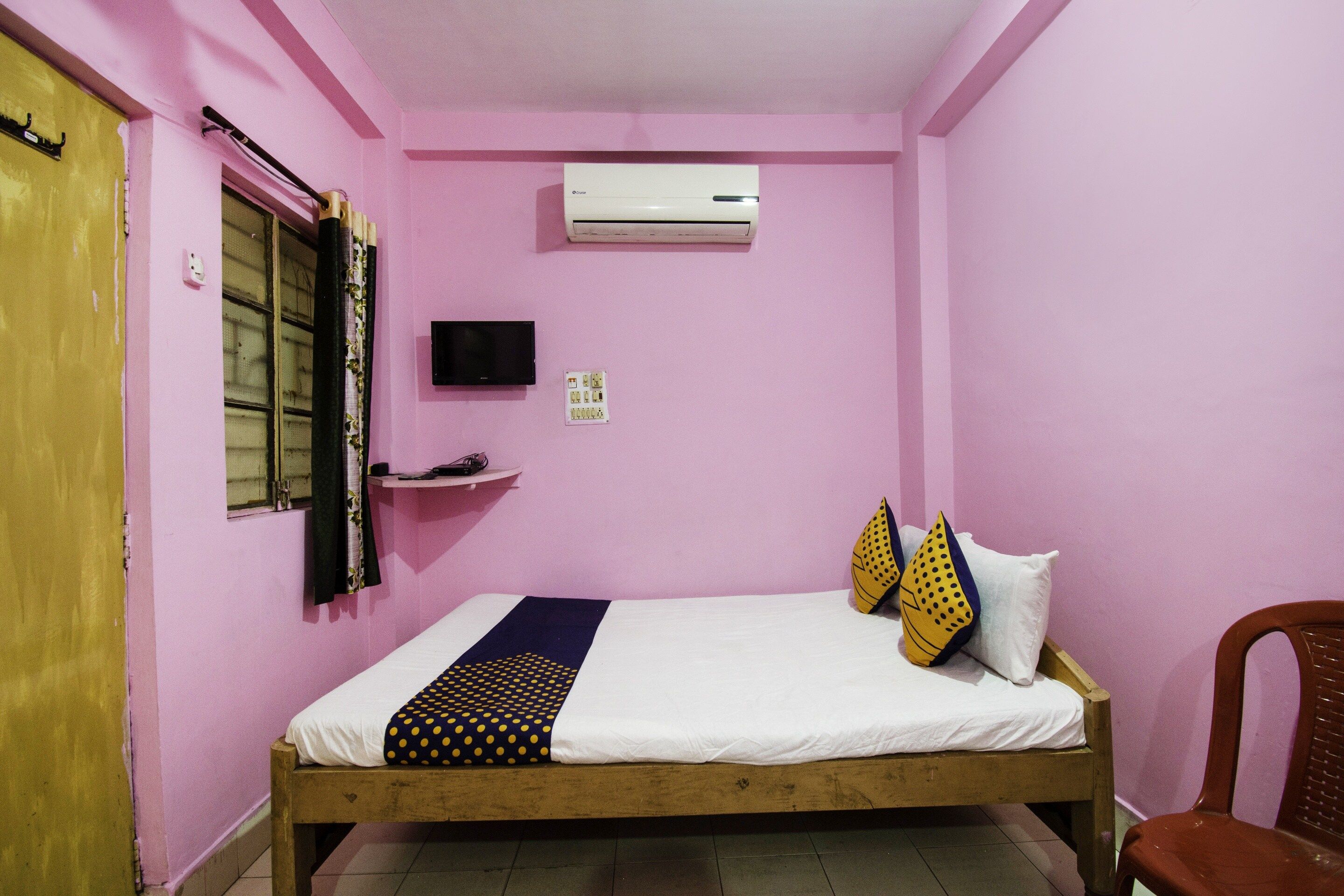 A single-bed room with a wooden frame, a white mattress, a black TV on the wall, a small shelf, and a chair.