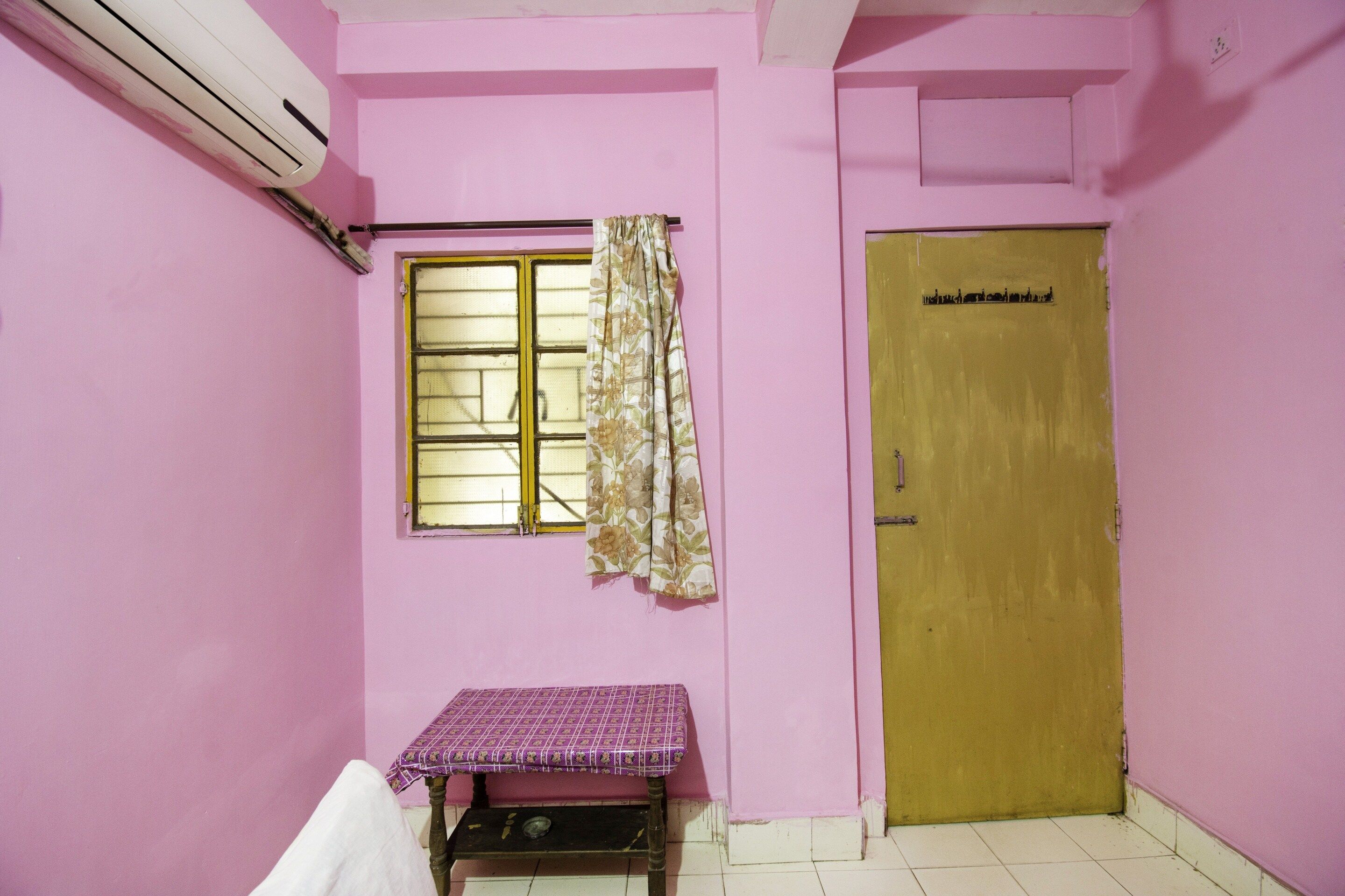 A room with pink walls, a window with a curtain, a wooden door, and a small table with a purple tablecloth.